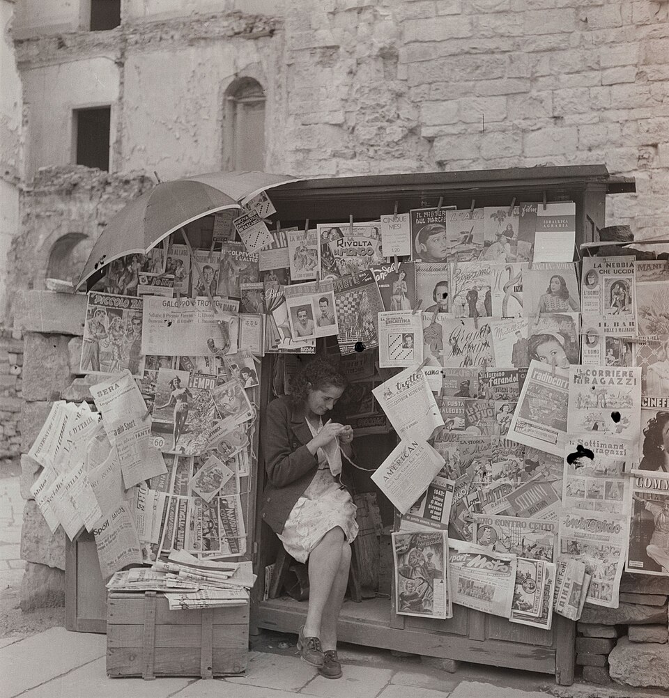 Una giornalaia lavora a maglia in un chiosco coperto di riviste, Roma 1947